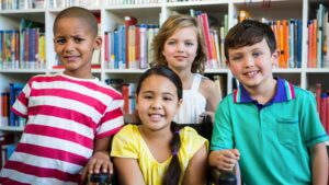 Four cheerful children smile together in a library classroom. One girl with long brown hair sits in a wheelchair. The children are posing in front of shelves filled with books. The sunny daytime photo would be great for projects about education, childhood, schools, and inclusion