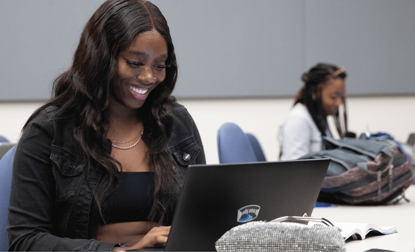 Student works on a laptop during class while another student studies in the background.