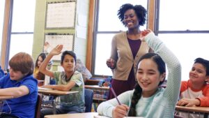 A teacher stands at the head of a classroom full of attentive students. Multiple children are raising their hands, hoping to answer the teacher's question.
