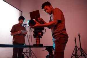 Two men are inside a studio space. One with curly hair and glasses holds a tablet, and the other adjusts a large video camera.