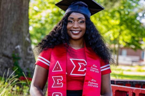 A female ECSU student wearing a graduation cap smiling at the camera with Greek stole and matching Greek affiliated shirt.