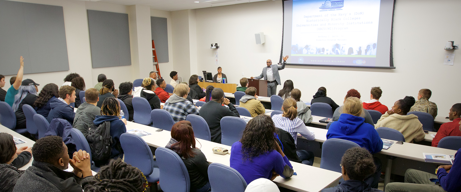 Workshop where a presentation is showing on a projector in a lecture hall with faculty seated in front of speaker.