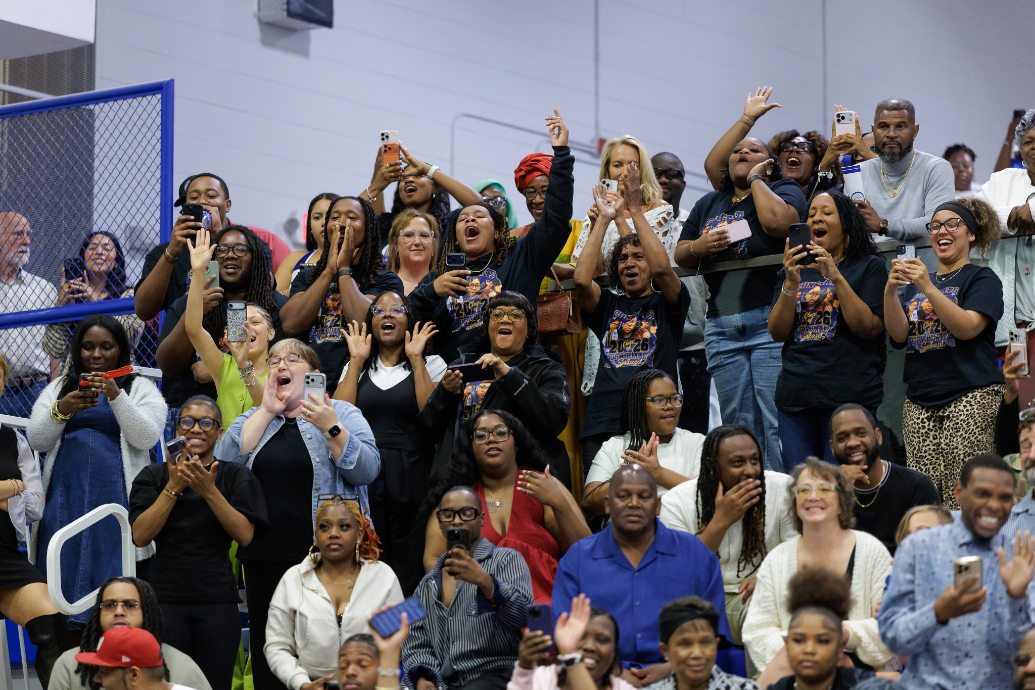 A large group of people seated in bleachers at an indoor ceremony cheer, clap, and hold up smartphones to record. Several people wear matching black T-shirts with a colorful graduation design that includes “Class of ’26,” while others wear casual clothing. A blue railing and gymnasium-style walls frame the background.