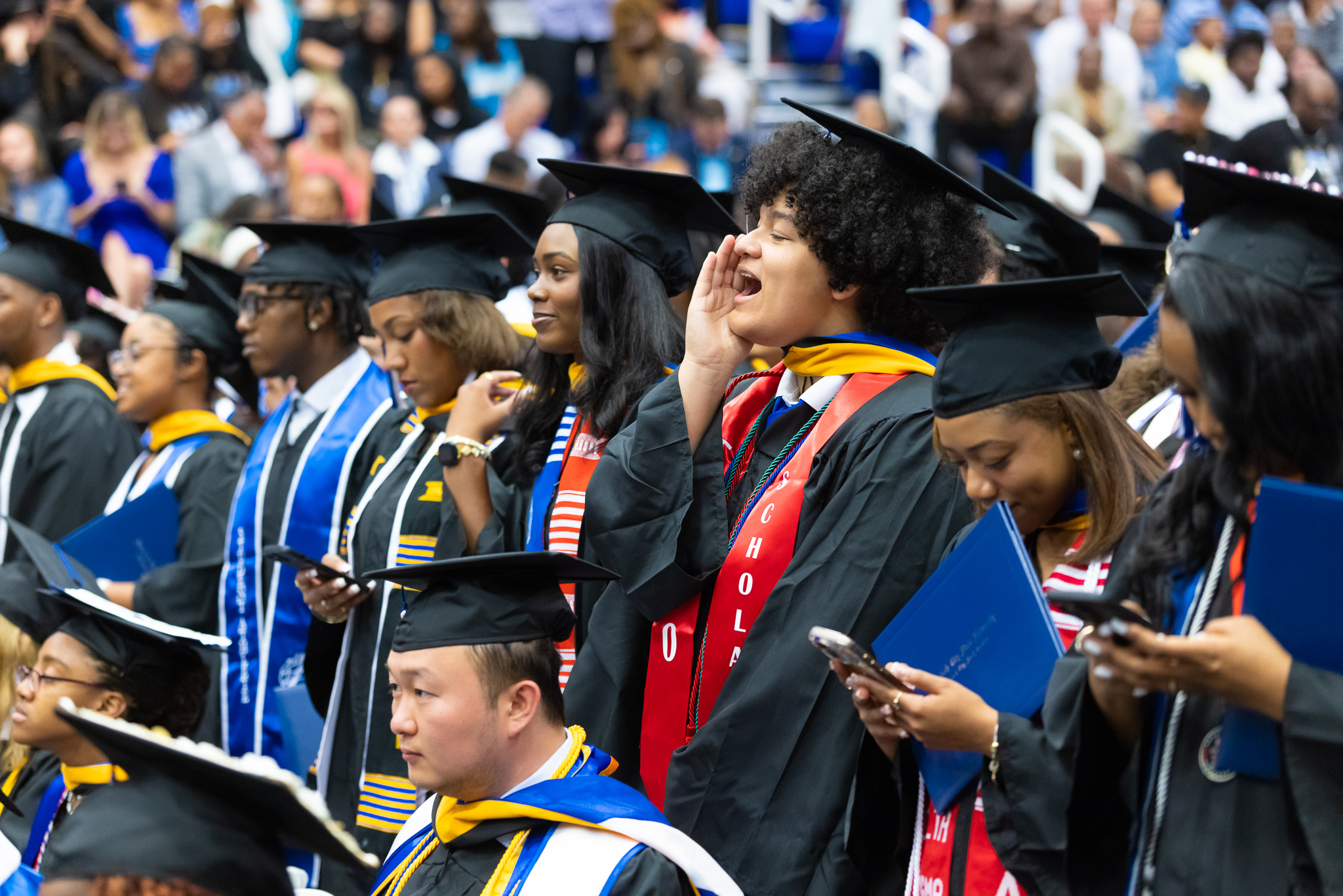 Row of graduates in caps and gowns seated during a commencement ceremony, wearing colorful stoles and cords. One graduate in the center cups a hand around their mouth as if calling out, while others hold programs, folders, or phones, with a large audience visible in the background.