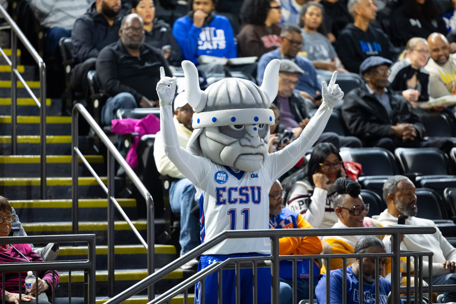 ECSU Viking mascot in a white and blue uniform stands in an indoor arena with arms raised, surrounded by spectators seated in the stands.