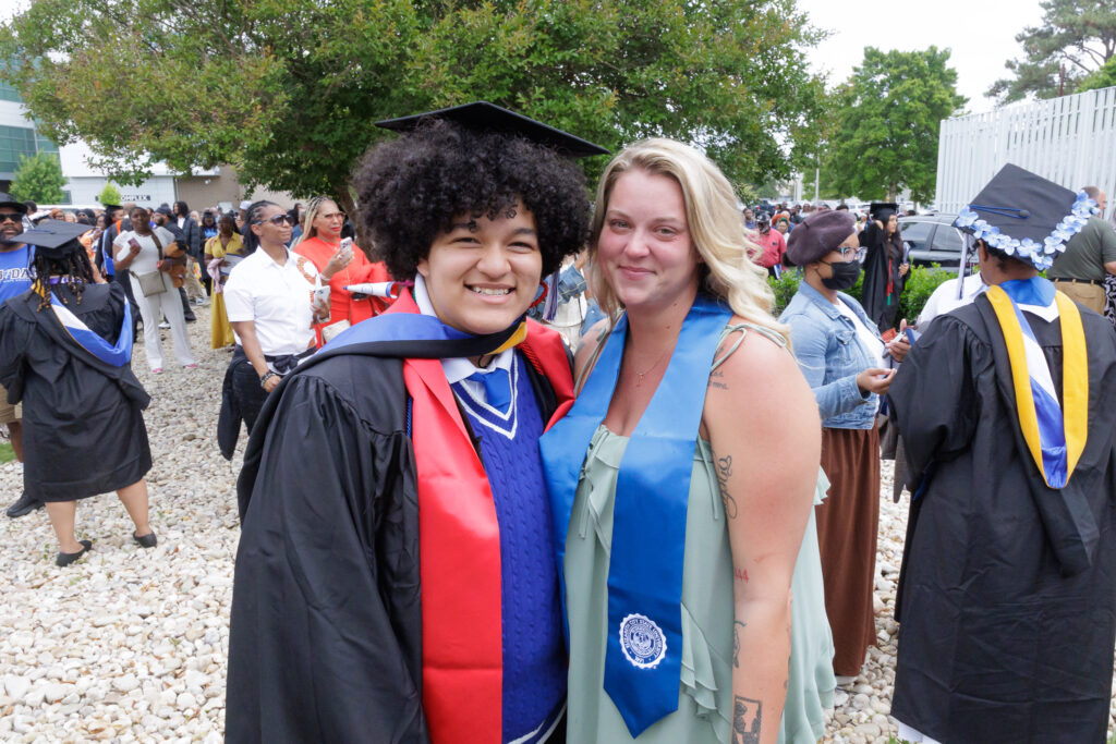 Two people pose together outdoors at a graduation celebration. One person wears a black cap and gown with a red and blue hood, while the other wears a light green dress with a blue stole featuring a university seal. A large crowd of graduates and attendees in caps and gowns fills the background near campus buildings and trees.