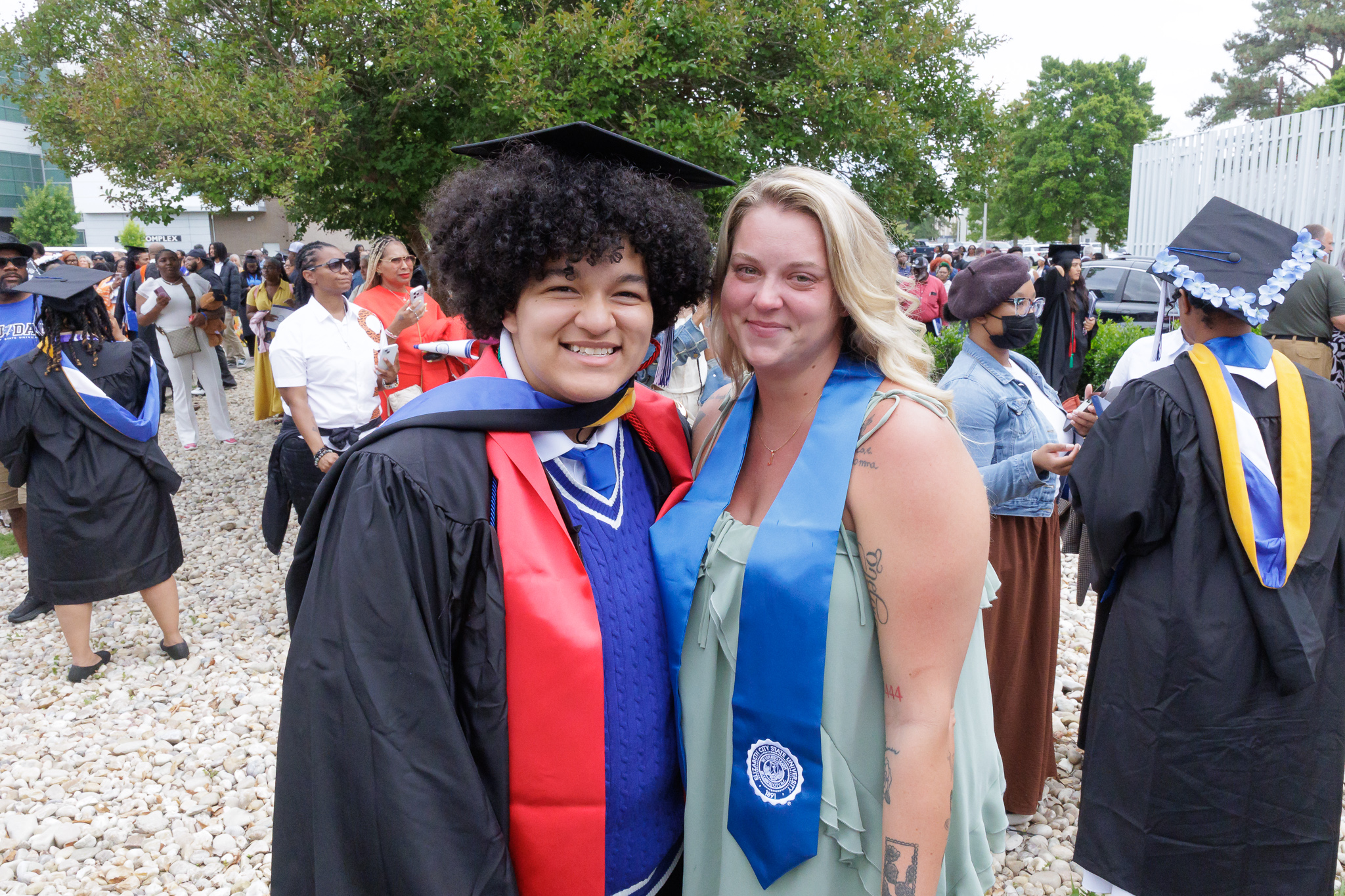 Two people pose together outdoors at a graduation celebration. One person wears a black cap and gown with a red and blue hood, while the other wears a light green dress with a blue stole featuring a university seal. A large crowd of graduates and attendees in caps and gowns fills the background near campus buildings and trees.