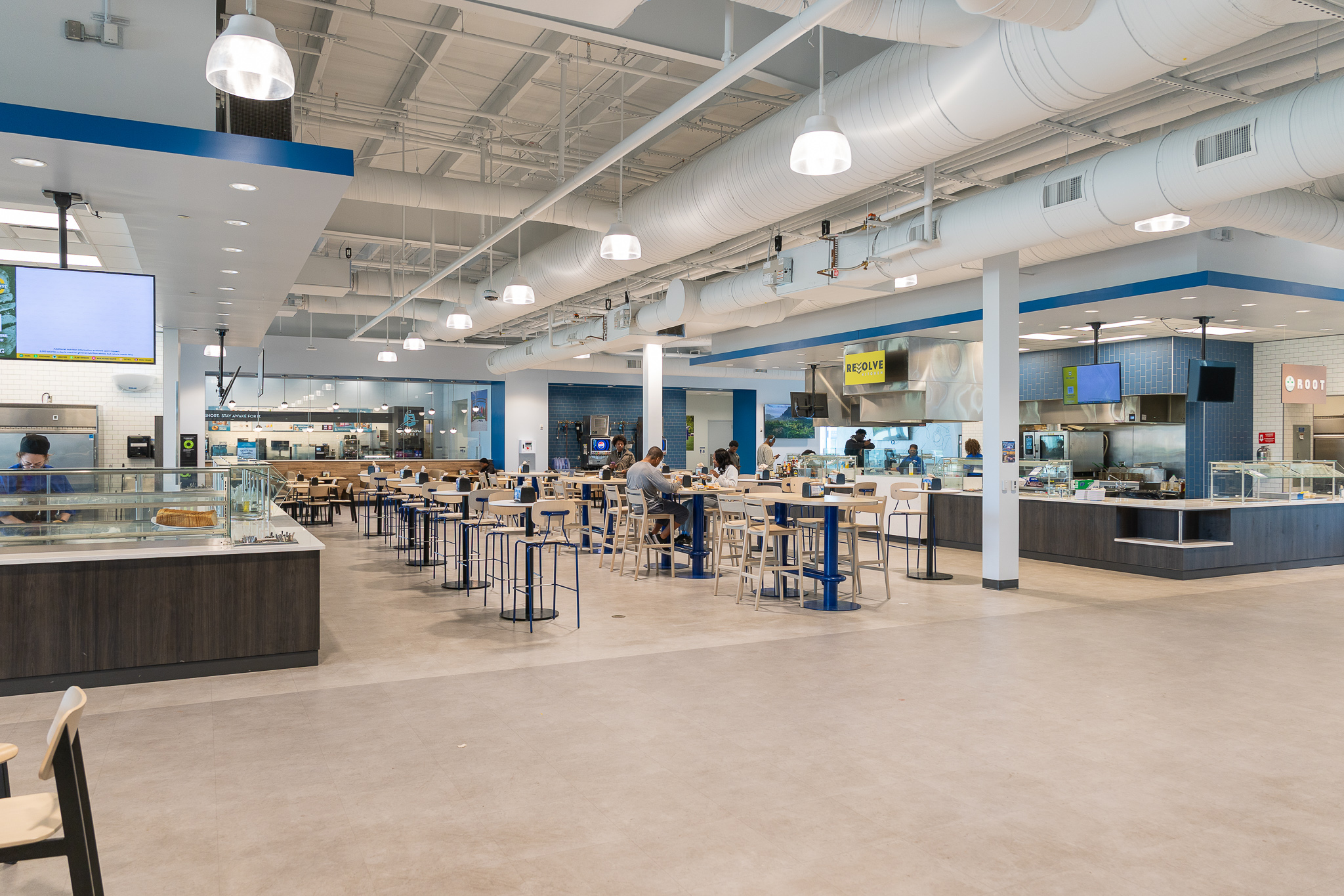 Interior of a modern campus dining hall with high ceilings and exposed ductwork, featuring tables, high-top seating, and multiple food stations. A few students sit and eat while service areas labeled “Resolve Kitchen” and “Root” are visible in the background.