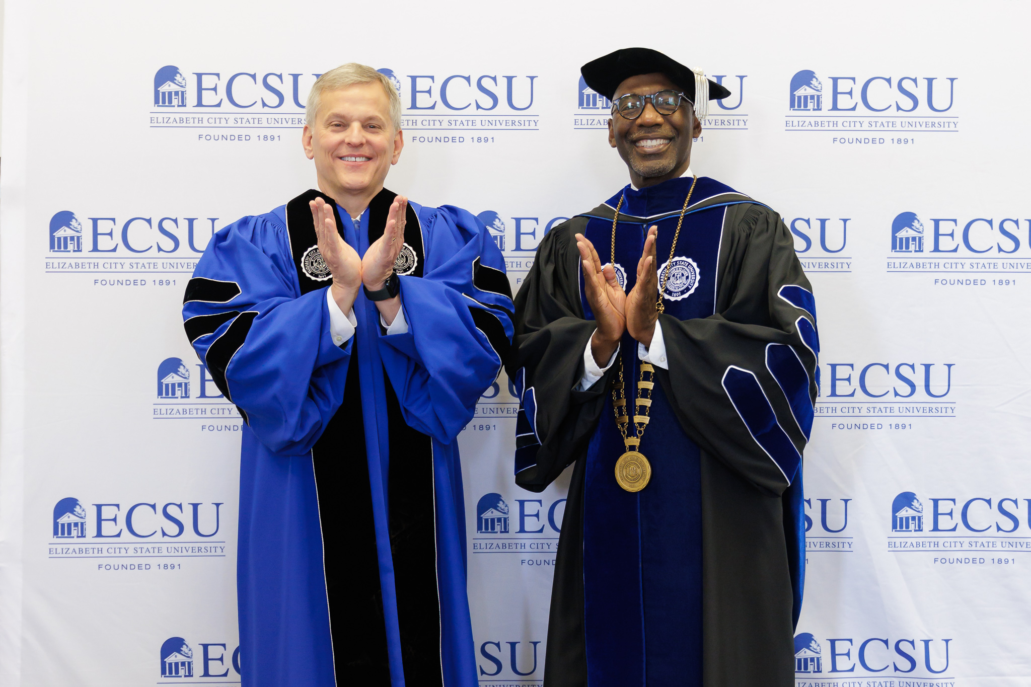 Two individuals in academic regalia stand side by side in front of a backdrop displaying repeated “ECSU – Elizabeth City State University, Founded 1891” logos. Both wear graduation gowns and clap with hands raised at chest level; one wears a blue robe, while the other wears a black robe with blue accents, a cap, and a ceremonial medallion.
