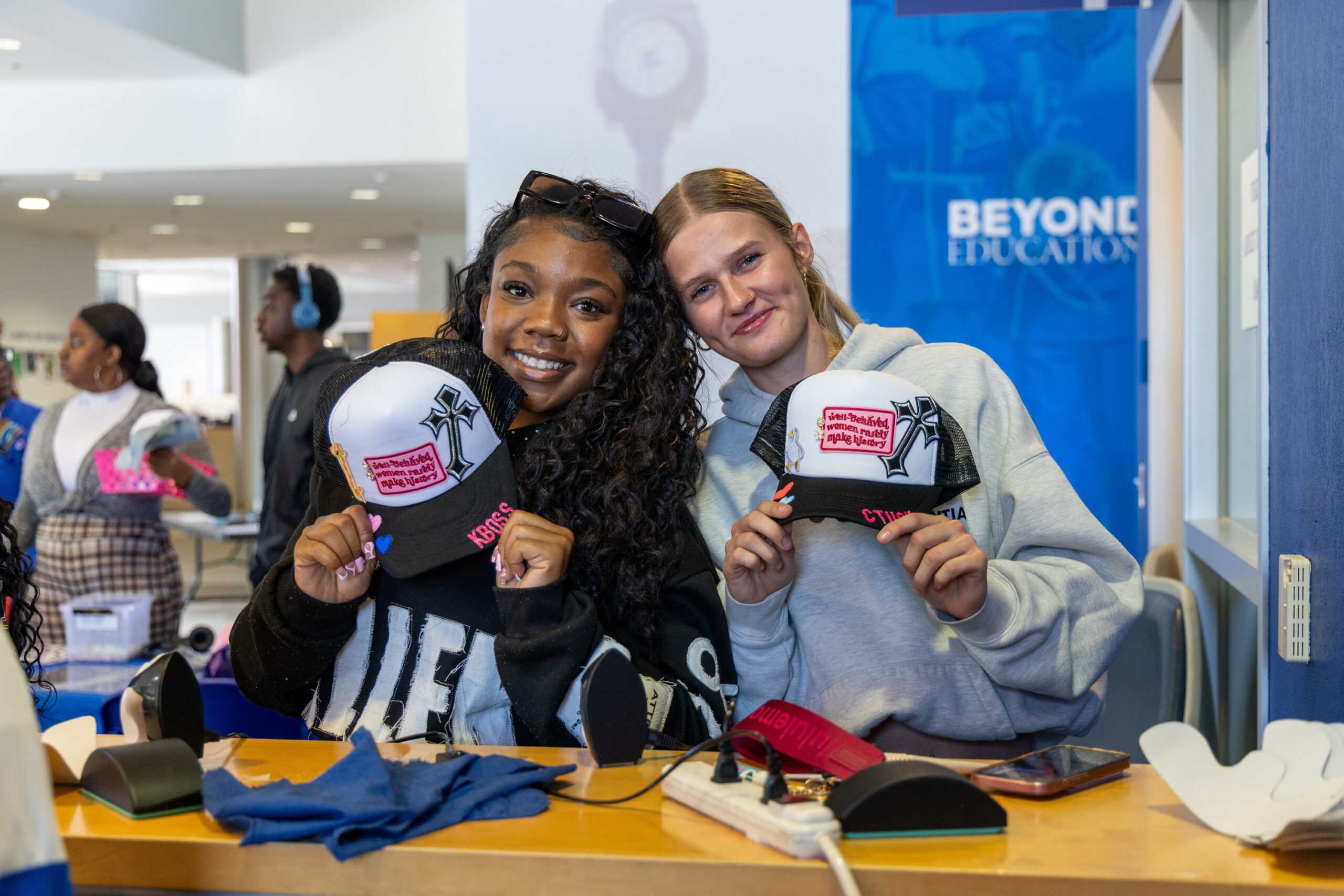 Two ECSU students smile and pose indoors while holding up decorated baseball caps with patches and designs. They stand at a table with crafting supplies, with other students and a banner reading “Beyond Education” visible in the background.