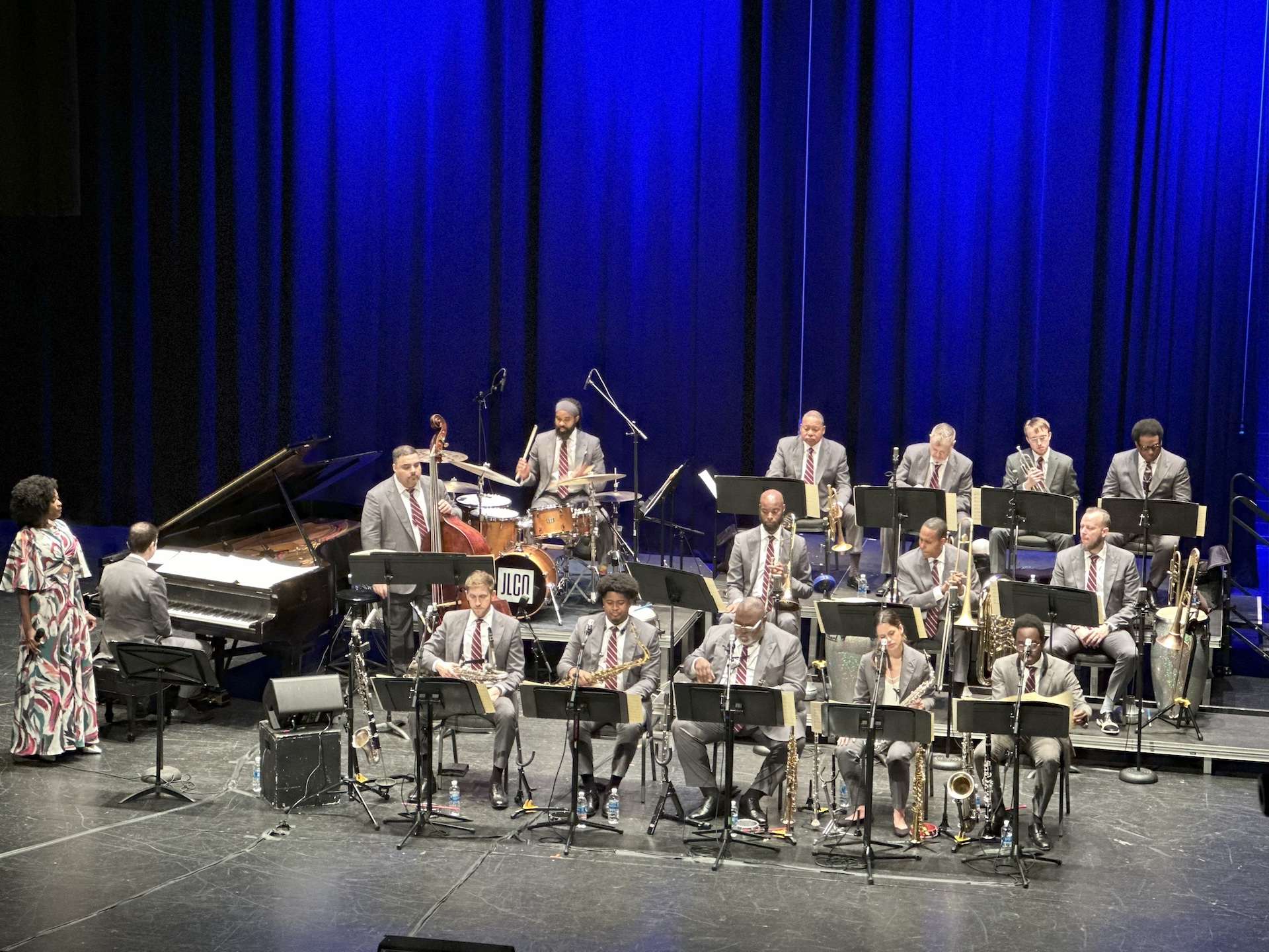 A large jazz ensemble performs on stage under blue lighting. Musicians seated in rows play instruments including saxophones, trombones, trumpet, bass, and drums, while a pianist and a standing vocalist appear at the left side of the stage. Music stands, microphones, and a raised platform for the rhythm section are visible.