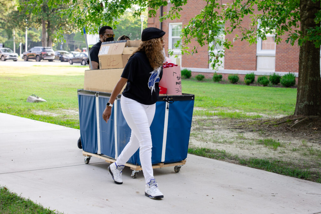 Two people move a cart loaded with boxes and belongings along a campus walkway, likely during move-in. One person walks in front while another pushes the cart, with trees and a brick campus building in the background.