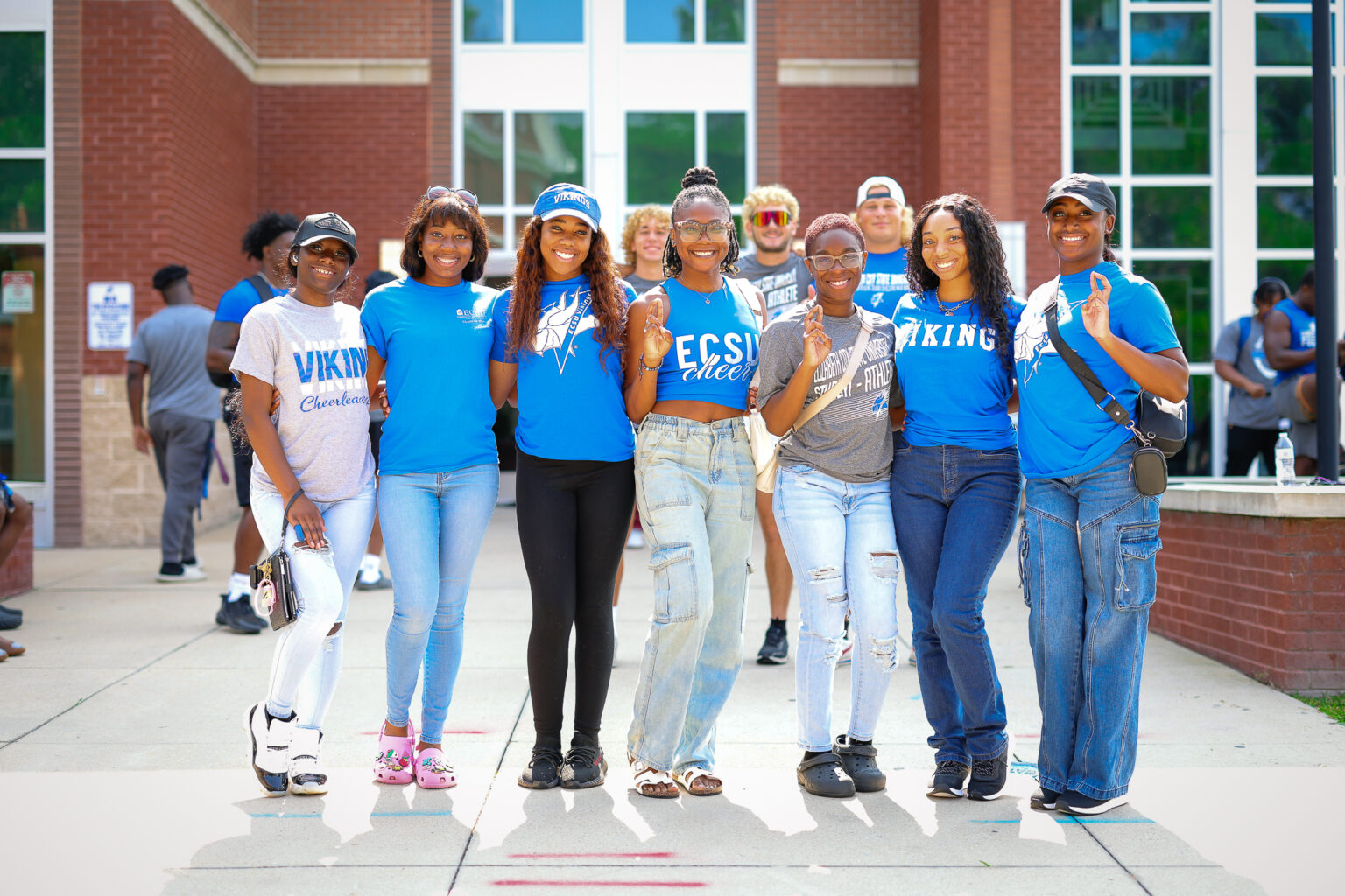Group of ECSU students stands together outside a residence hall, smiling and posing with arms around each other. Many wear blue shirts with Viking or ECSU logos, creating a strong school spirit atmosphere.