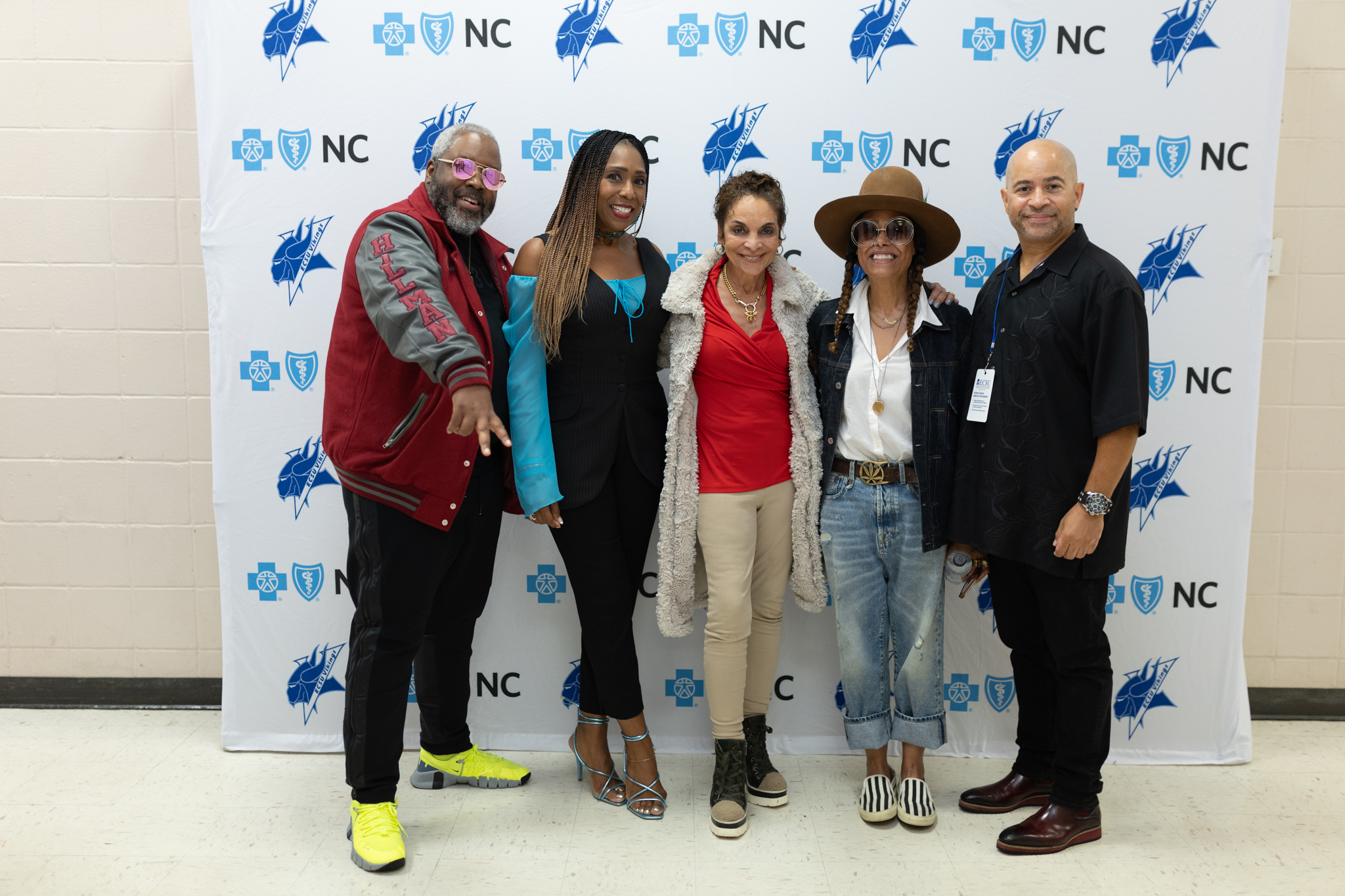 Six people pose together in front of a step-and-repeat backdrop displaying multiple logos, including “NC” and health-related symbols. The group stands side by side indoors, wearing a mix of casual and dressed-up outfits, including a red jacket, a black outfit with blue accents, a red top with a light-colored coat, and a wide-brim hat. One person gestures toward the camera, while others stand close together for the photo.