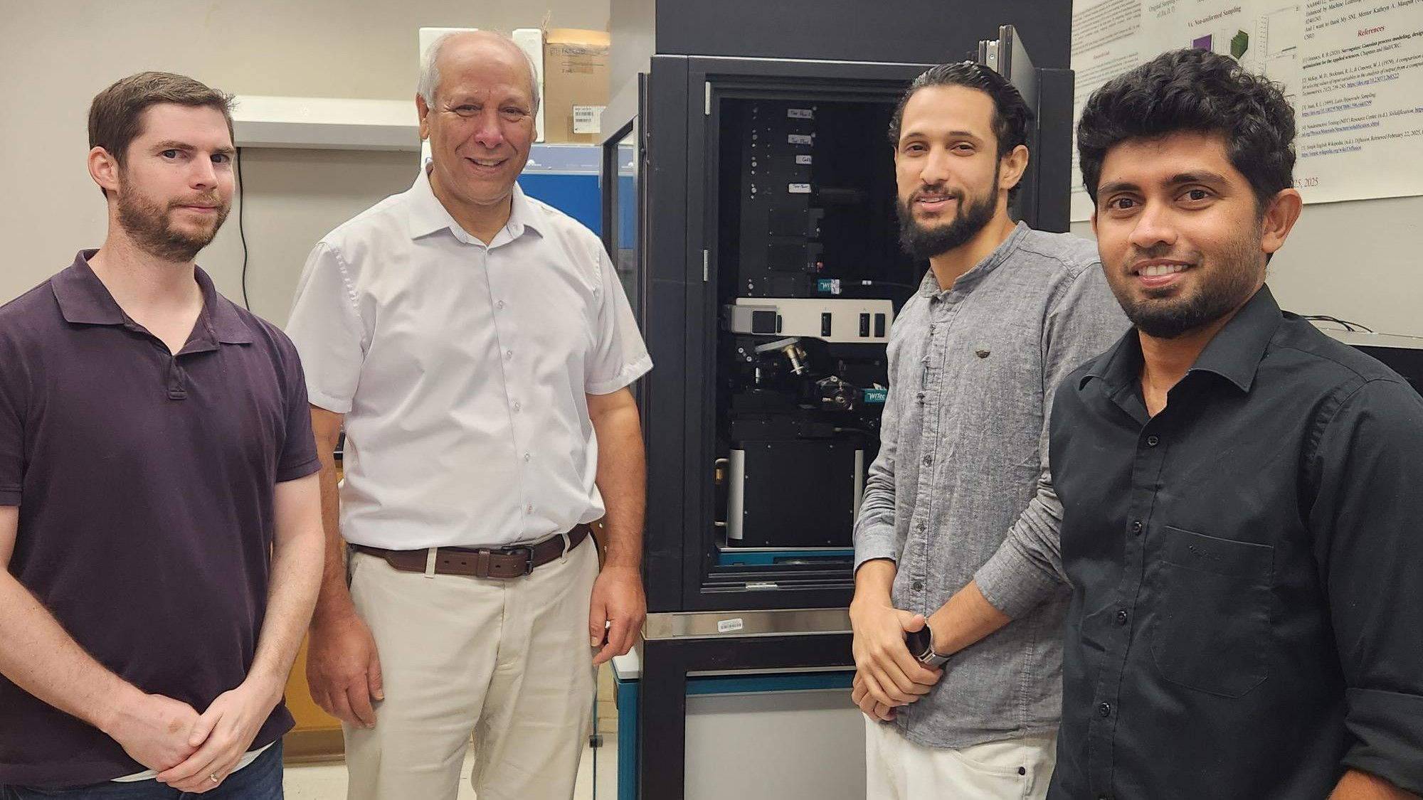 Four researchers stand in a laboratory beside a large scientific instrument, posing for a group photo in a research environment with equipment and lab materials in the background.