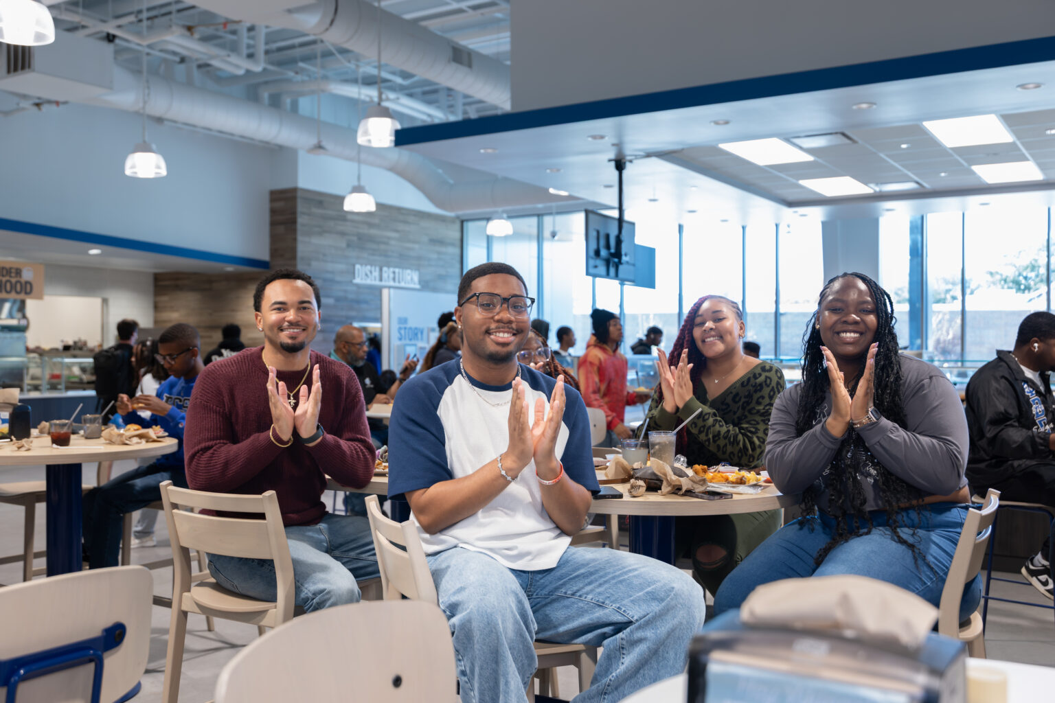 Four students sit at a table inside a campus dining hall, clapping while facing the camera. The space features bright lighting, large windows, and overhead fixtures, with other students eating and socializing in the background near food stations labeled “Dish Return.”