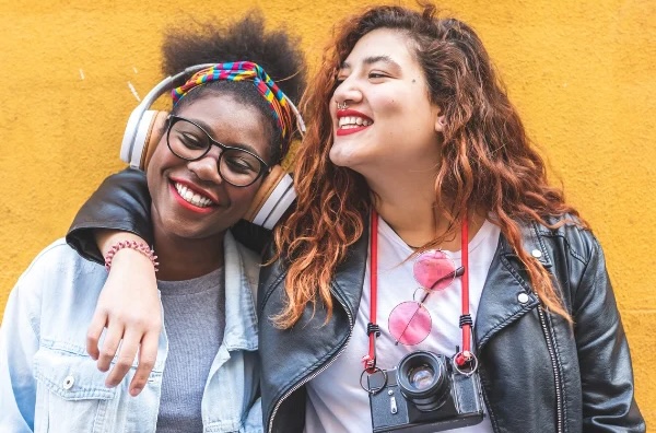 Two friends stand close together against a bright yellow wall, smiling and laughing. One wears glasses and headphones while the other has a camera hanging around her neck; they lean into each other with an arm draped over the shoulder, showing a joyful, candid moment.
