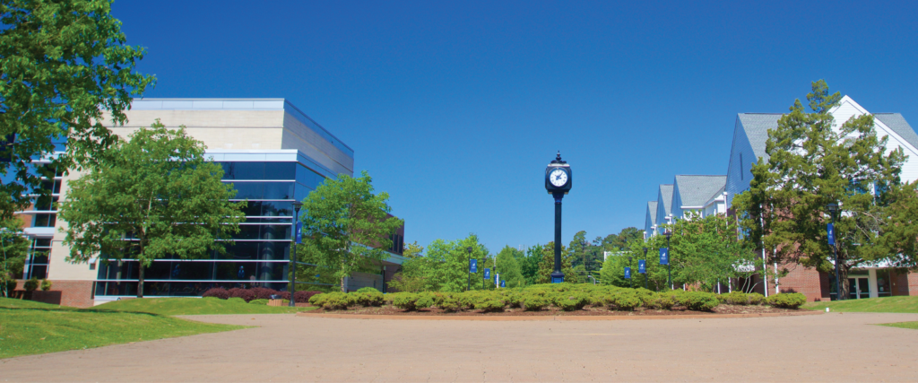 Wide view of a university campus walkway under a clear blue sky, featuring a landscaped circular area with a tall clock tower at the center. A modern building with large glass windows stands on the left, while residential-style campus buildings line the right side. Trees, grass, and campus banners line the path, creating a symmetrical, open outdoor setting.