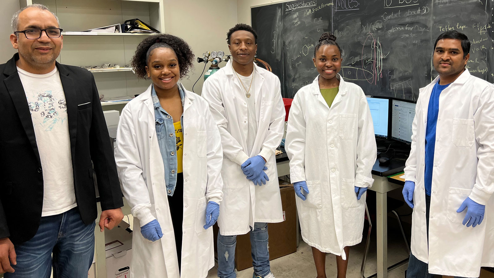 Five students and a professor stand together in a laboratory wearing white lab coats and gloves, smiling in front of a chalkboard covered with scientific notes and diagrams.