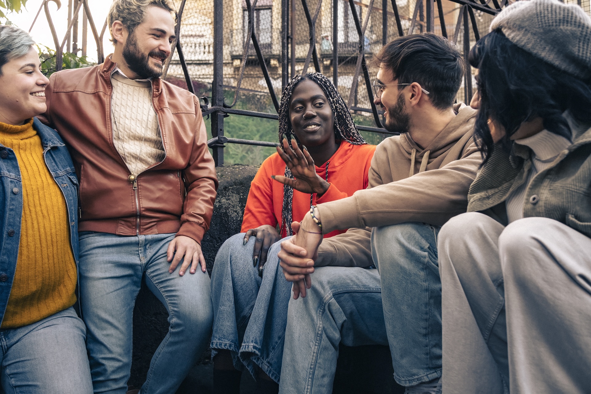 Five friends, a mix of women and men, sit outdoors in the daytime sunlight chatting and enjoying each other's company.