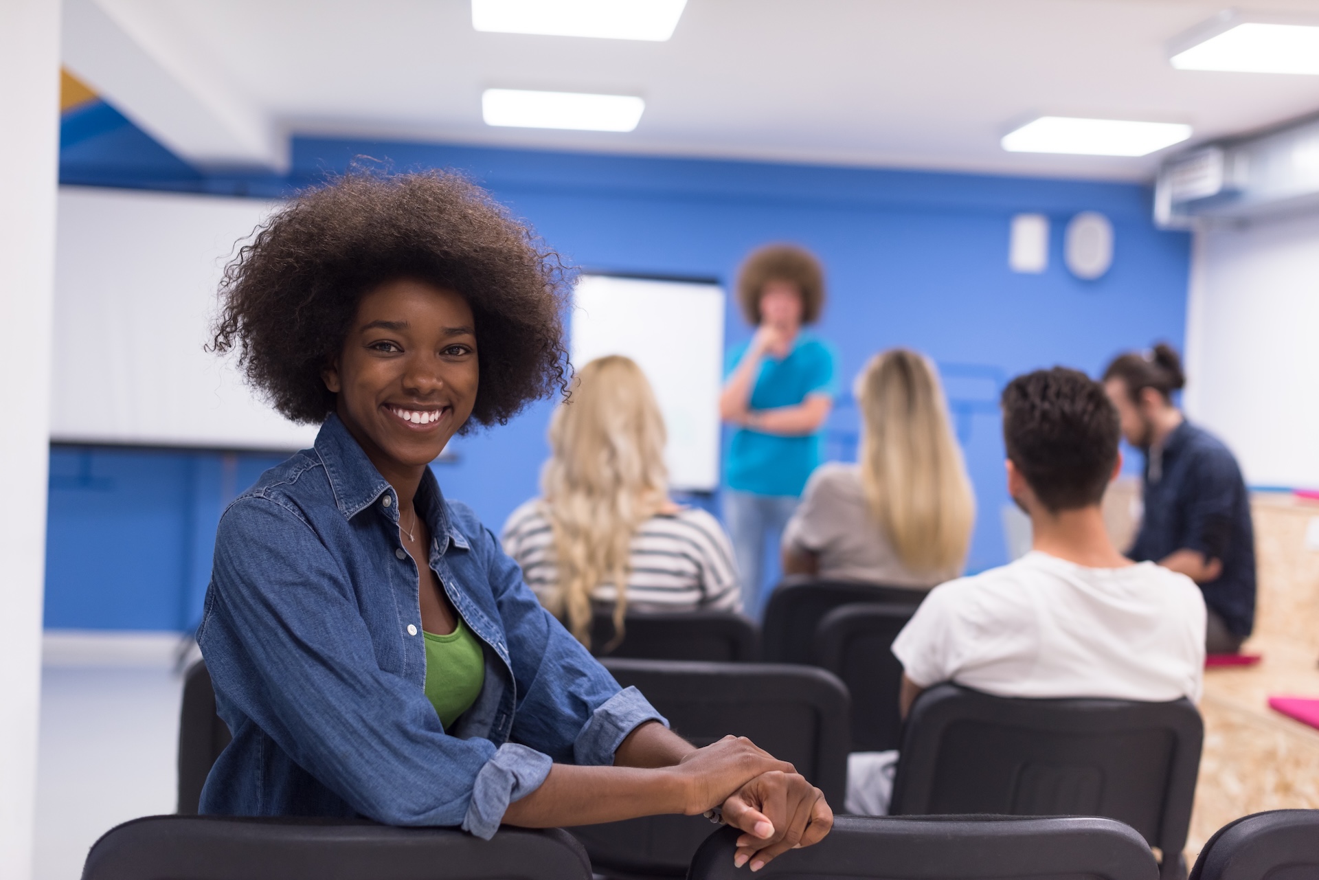 A smiling woman in a denim jacket sits in the foreground of a bright, modern office space, where several people are seated in rows of black chairs,