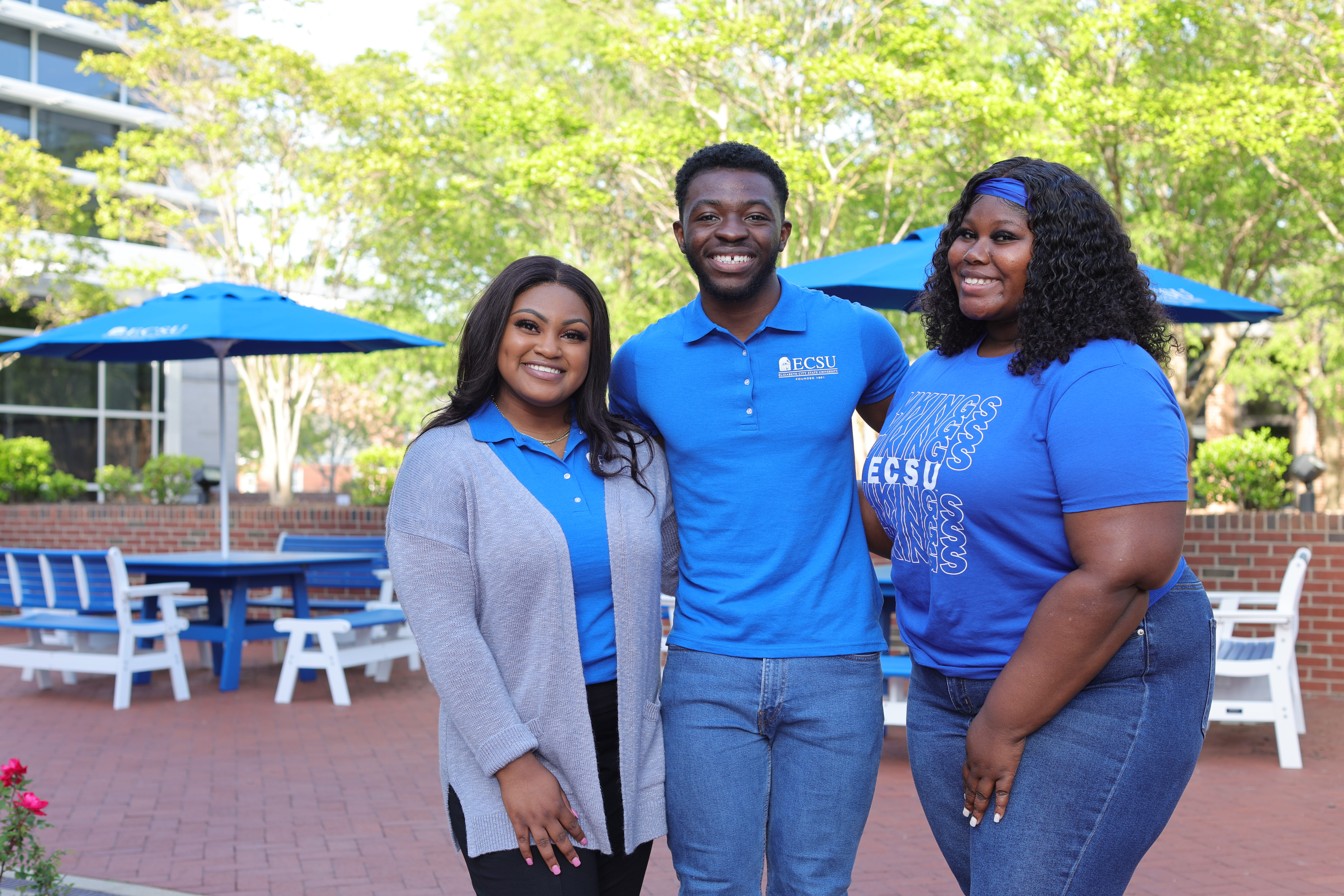 Three ECSU students stand together smiling in an outdoor campus courtyard, wearing blue shirts with ECSU branding. Blue tables and umbrellas, brick walkways, and trees are visible in the background, creating a bright, welcoming campus setting.