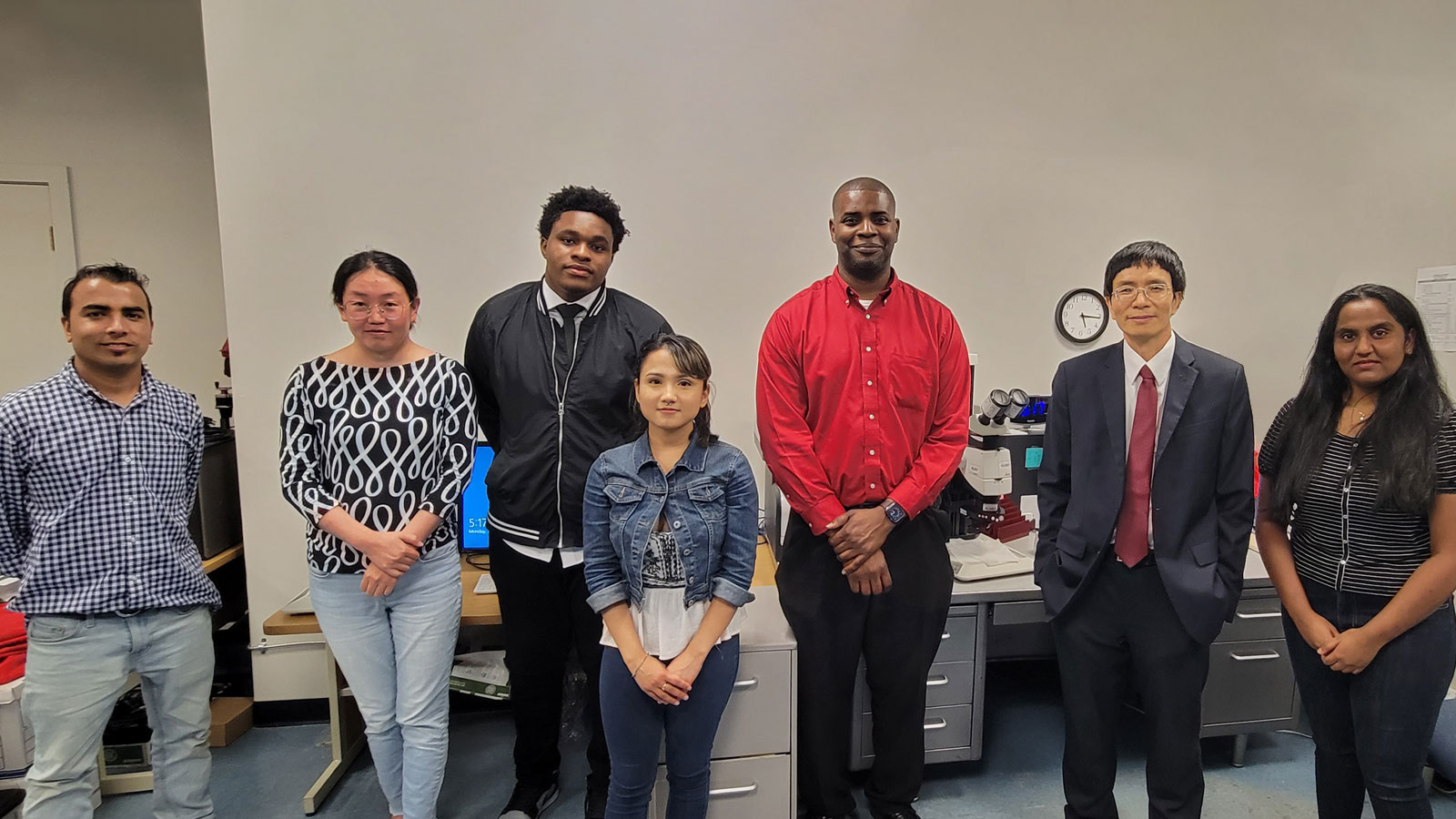 Group of students and faculty stand together in a research lab, posing in front of computer workstations and scientific equipment in a classroom setting.