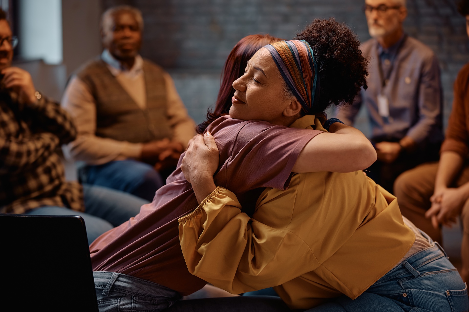 In a dimly lit interior setting, a group of adults participate in a therapy session. A woman with curly hair and a colorful headband tightly embraces another woman with reddish-brown hair.