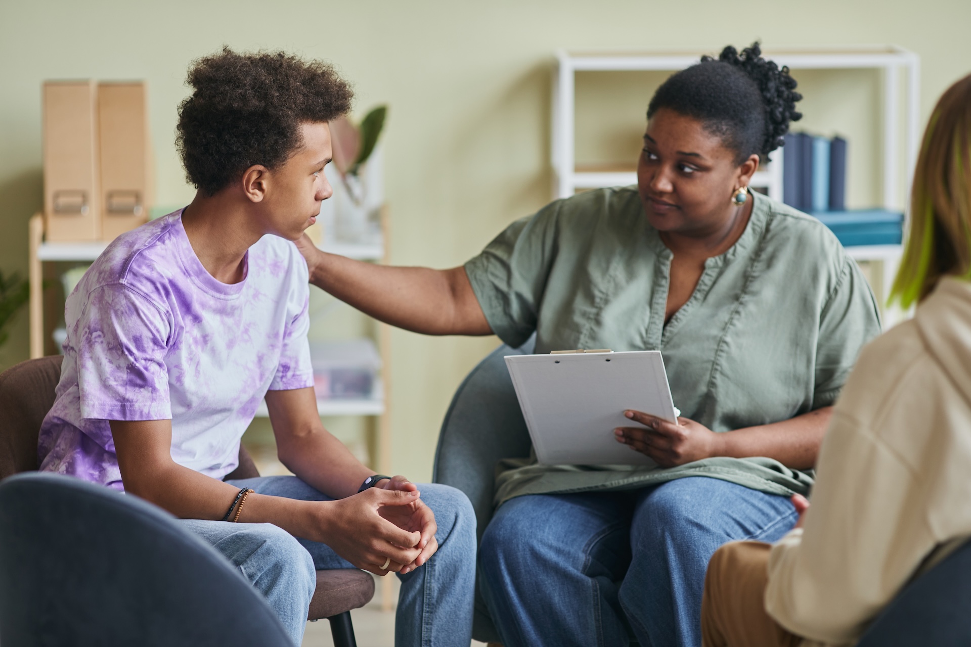 A teen with curly dark hair sits in a chair wearing a purple tie dye shirt with blue jeans. An adult female with dark hair in a bun wearing an olive green blouse and blue jeans is holding a notepad, with a hand on the teen's shoulder.