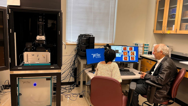 Student and instructor sit at a computer workstation in a laboratory while reviewing imaging data next to a large scientific instrument, with additional equipment and cables visible around the workspace.