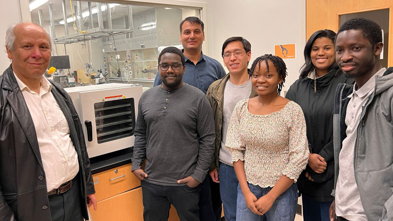 Group of students and faculty pose together inside a laboratory, standing in front of equipment and glass windows that look into an adjacent lab space.