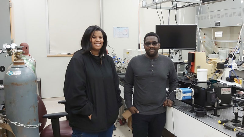 Two students stand in a laboratory next to scientific equipment and gas tanks, smiling at the camera in a research workspace filled with instruments and monitors.
