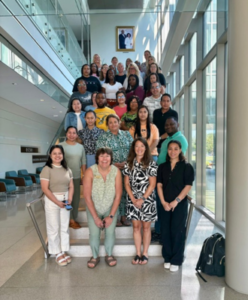 Large group of students and faculty pose together on an indoor staircase in a bright building with tall windows, smiling for a group photo.